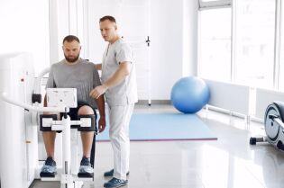 Grafika przedstawia dwóch mężczyzn, z których jeden jest pacjentem ćwiczącym na urządzeniu do rehabilitacji, a drugi go nadzoruje.