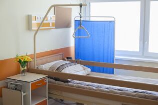 Grafika przedstawia pokój w hospicjum z widocznym łóżkiem oraz szafką na podręczne rzeczy.